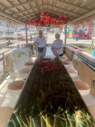 Big Kahuna Yacht Photos Pics 2022 Beachcat COI Tiki Boat with two people onboard, featuring a long table and tropical decor.