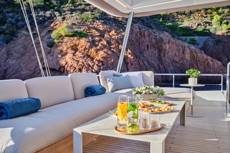  Yacht Photos Pics Luxurious 2023 Lagoon Seventy 7 yacht deck with elegant seating and refreshments, scenic coastal backdrop.