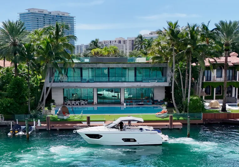  Yacht Photos Pics Luxury yacht cruising by modern waterfront mansion with pool, surrounded by palm trees.