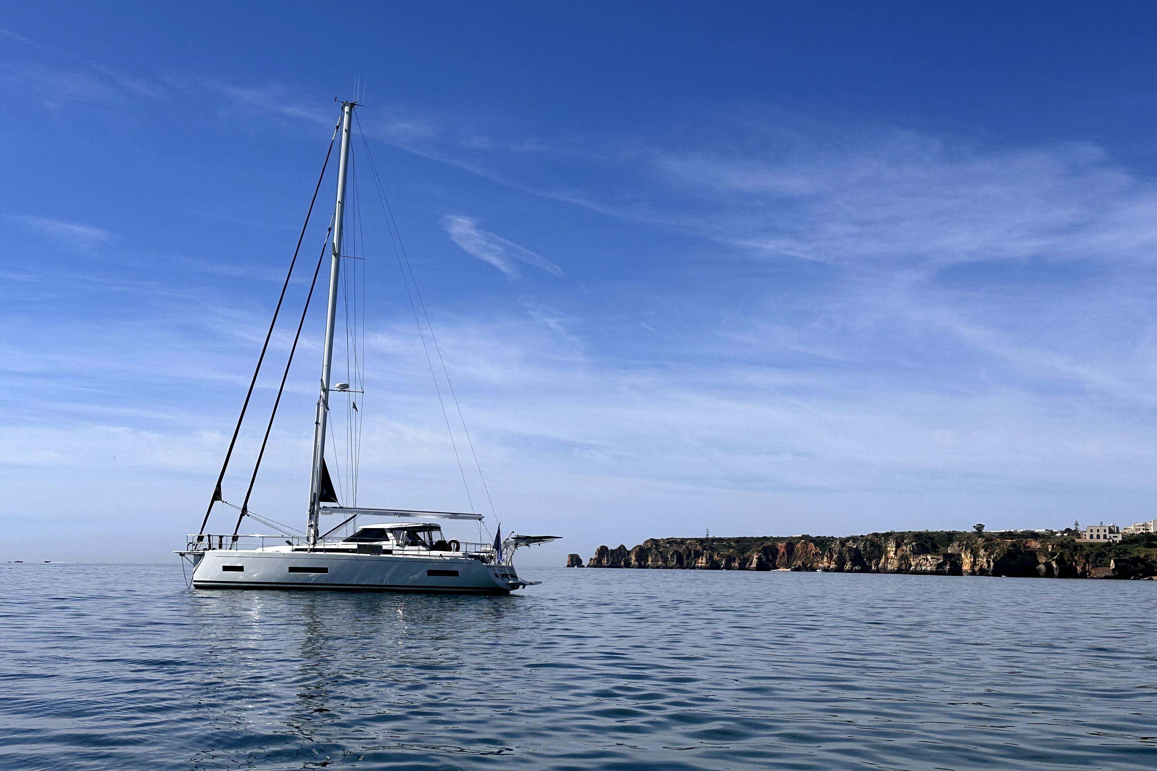 2024 Amel 50 yacht sailing near rocky coastline under clear blue sky.