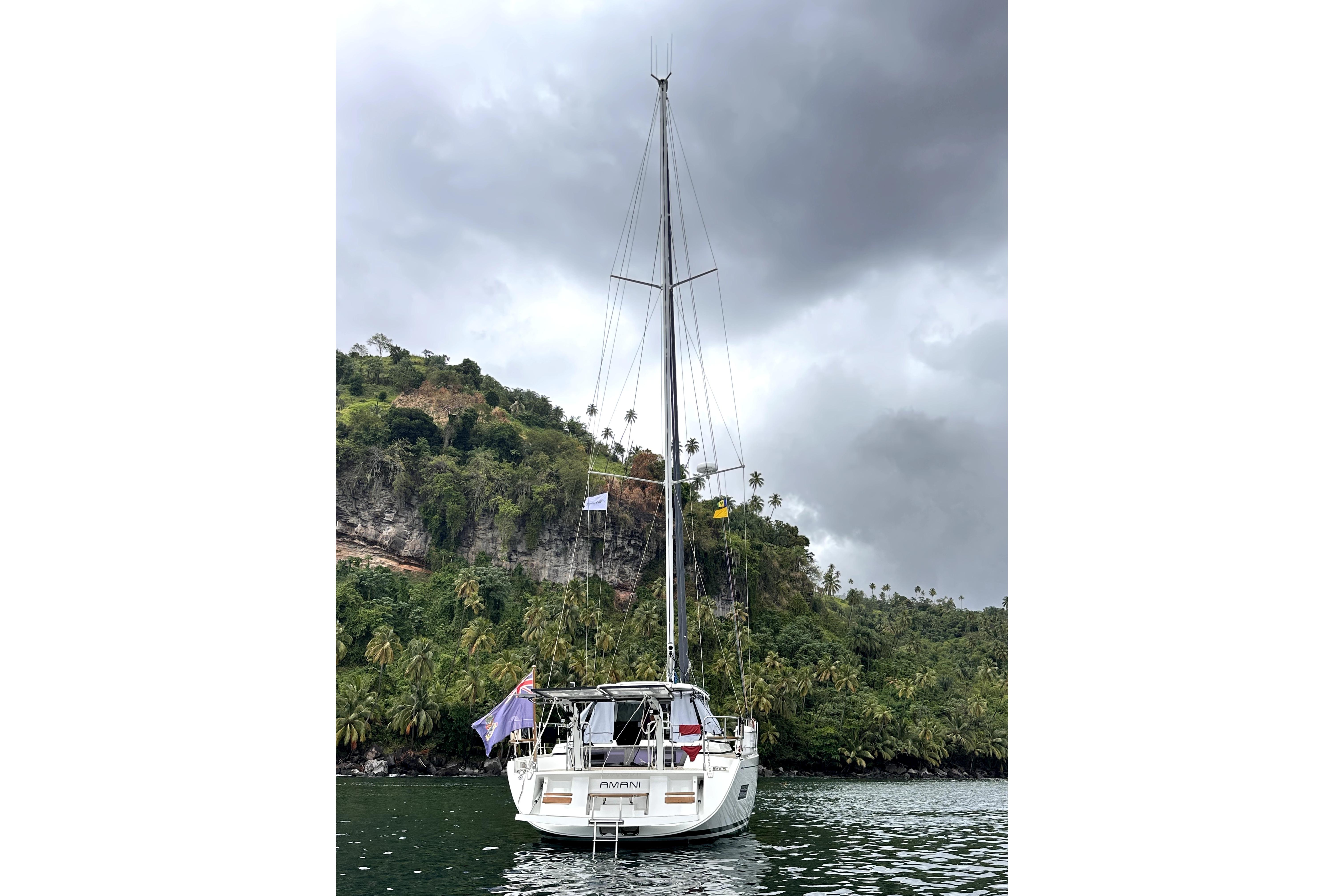 2024 Amel 50 sailboat anchored near lush, tropical coastline under cloudy skies.