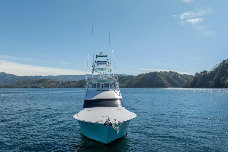 Scandaloso Yacht Photos Pics Front view of a 2006 Viking 56 yacht on calm waters, surrounded by scenic hills.