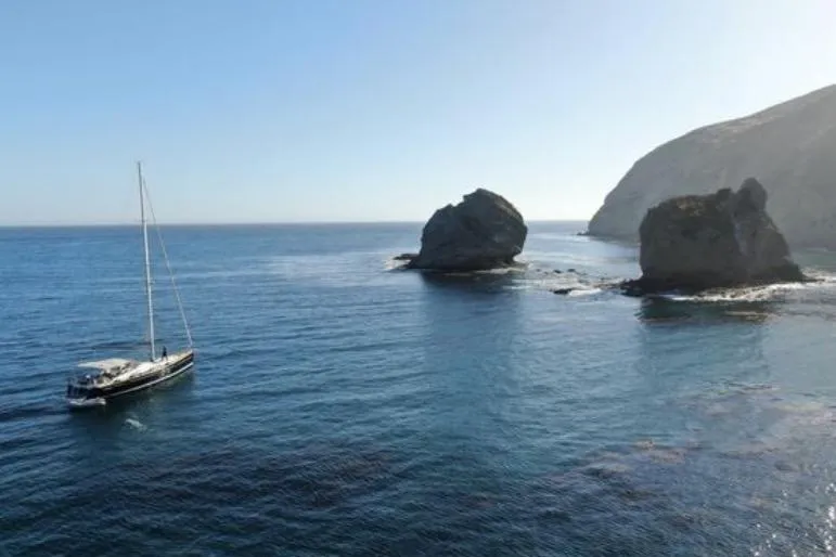 Motu Yacht Photos Pics Sailing yacht Jeanneau 57 (2013) near rocky coastal landscape under clear blue sky.