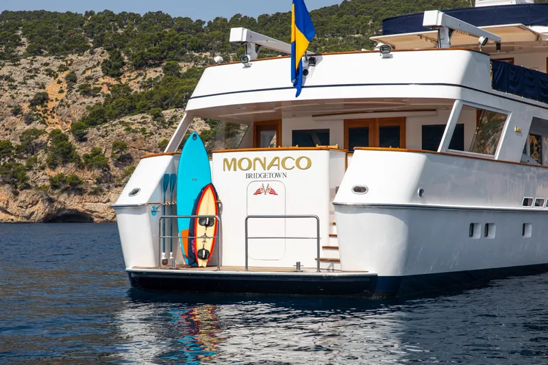 Monaco Yacht Photos Pics Luxury 1982 Feadship Classic yacht "Monaco" with surfboards, anchored near scenic cliffs.