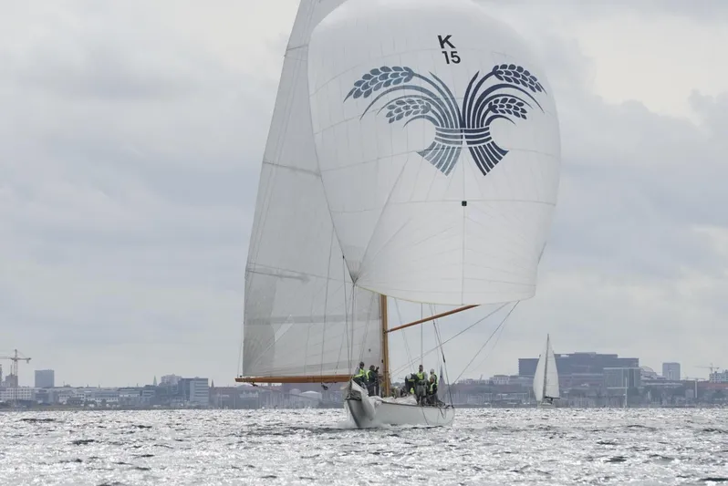 Wings K-15 Yacht Photos Pics Classic 1937 12-metre sailboat with spinnaker, sailing on a cloudy day near city skyline.