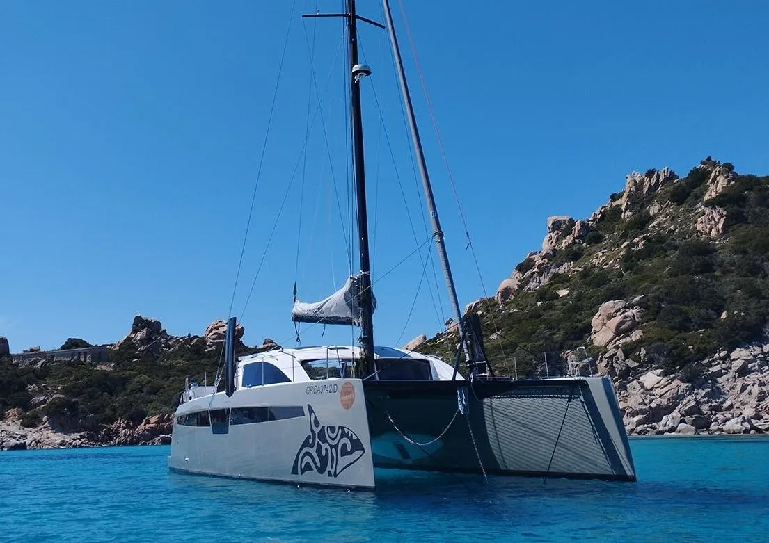 2025 Comar C-Cat 48 catamaran sailing in clear blue waters near rocky coastline.
