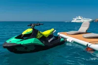 Jet ski docked near floating platform with lounge chairs, yacht in background, clear blue sea.