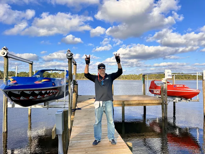 Scream Machine Yacht Photos Pics Man celebrating on dock with two colorful jet boats, under a bright blue sky.