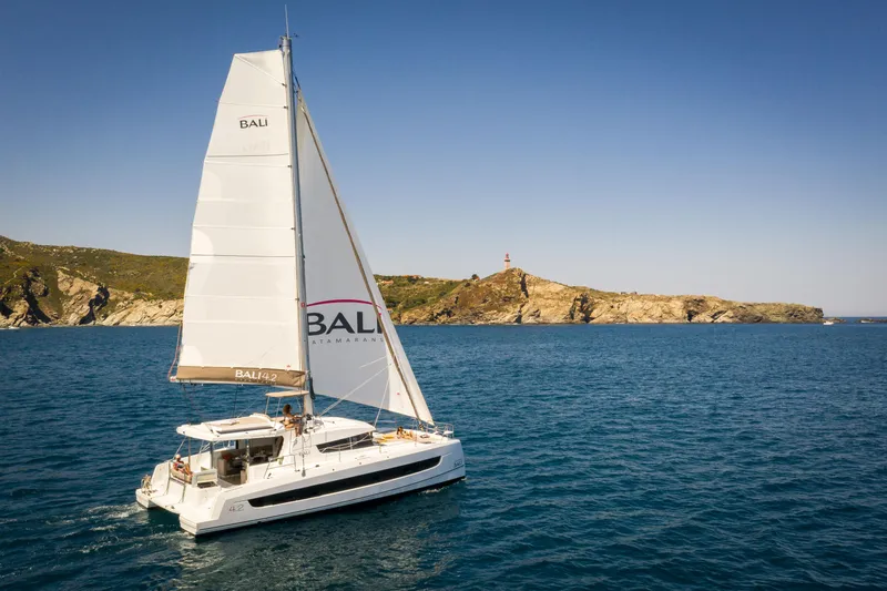 Bali 4.2 Yacht Photos Pics Sailing catamaran Bali 4.2, 2026 model, cruising near rocky coastline under clear blue sky.