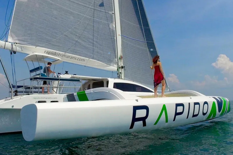  Yacht Photos Pics 2026 Rapido 60 trimaran sailing with people on deck, clear skies, and calm waters.