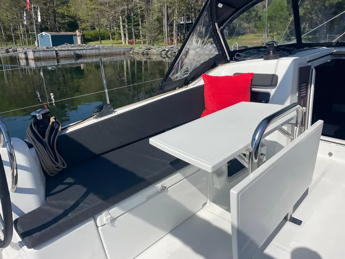  Yacht Photos Pics 2024 Jeanneau Sun Odyssey 380 cockpit with seating, table, and red cushion, docked by a serene lake.