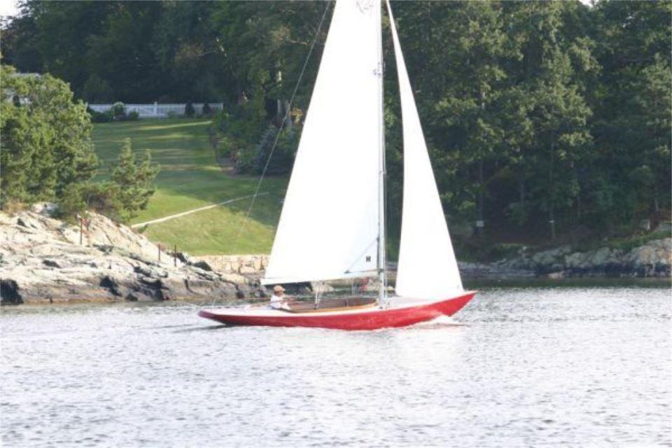 Red Rustler 24 sailboat from 2025 sailing near a rocky shoreline with lush greenery.