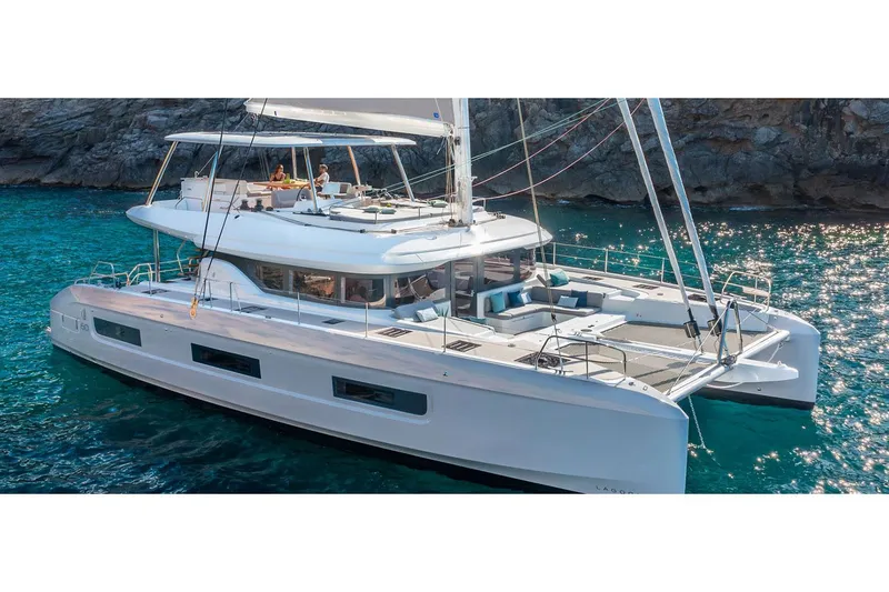  Yacht Photos Pics Manufacturer Provided Image: 2025 Lagoon 60 catamaran sailing in clear blue waters near rocky coastline.