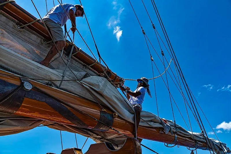 Invader Yacht Photos Pics Two people working on the sails of a 1905 Lawley Schooner under a clear blue sky.