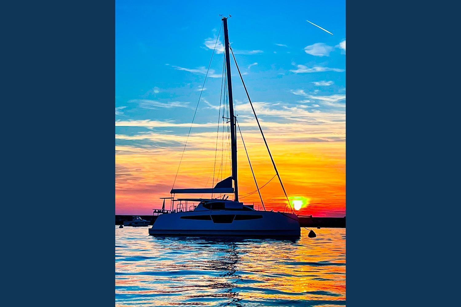 Sailing catamaran Comar C-CAT 37 at sunset, vibrant sky reflecting on water.
