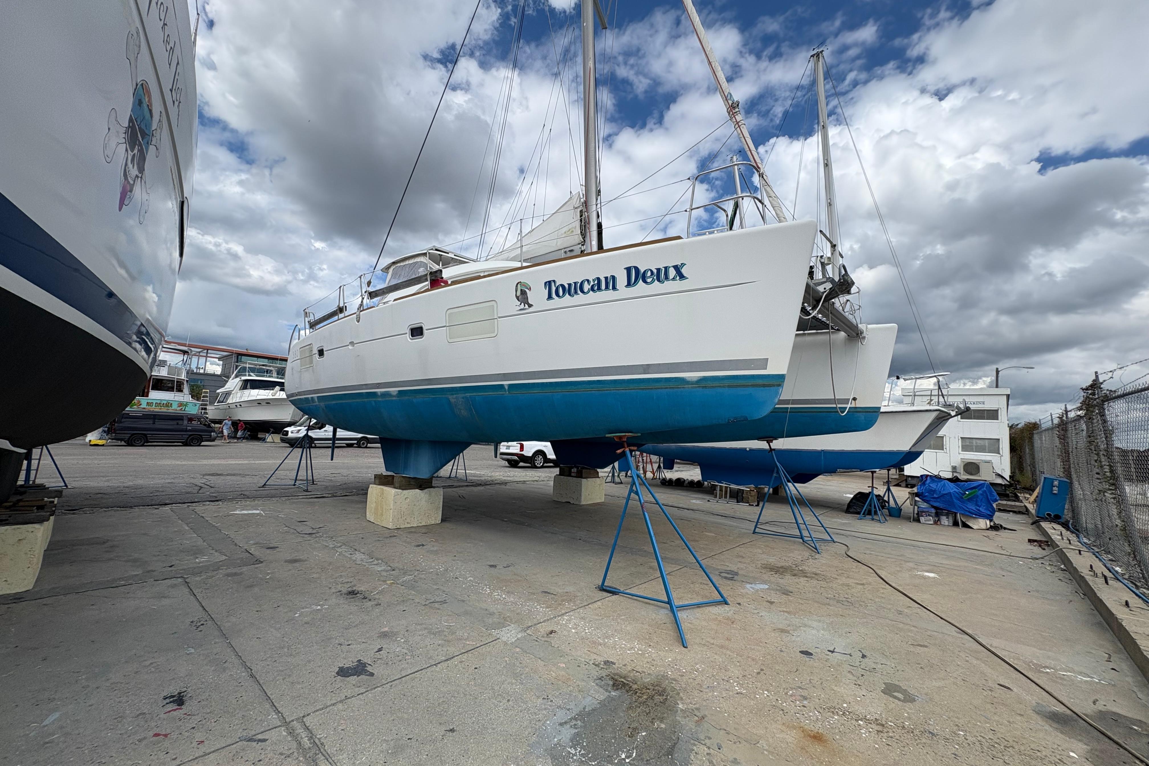 2004 Lagoon 380 catamaran "Toucan Deux" on stands in a boatyard under cloudy skies.